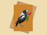 A greeting card with a warm mustard-orange background, featuring an illustration of a black-and-white bird holding red flowers, shown with a kraft envelope behind it on a light beige background.