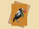 A greeting card with a warm mustard-orange background, featuring an illustration of a black-and-white bird holding red flowers, shown with a kraft envelope behind it on a light beige background.