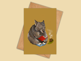 A greeting card with a warm mustard-yellow background, featuring an illustration of a wombat reading a red book next to a small green plant. The card is displayed with a brown kraft envelope behind it on a solid light beige background.