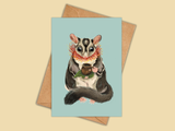 Card with a sugar glider illustration holding a cup on a beige background