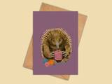 A rectangular card with a purple background featuring an illustration of an echidna holding knitting needles and yarn, shown with a brown envelope on a warm beige background.