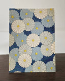 A square fabric or paper art print featuring a repeating pattern of large daisy-like flowers in cream, light blue, and navy blue with yellow centers, standing on a dark wooden surface against a white wall.