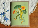 Framed artwork of a frog among flowers on a wooden surface with lace fabric.