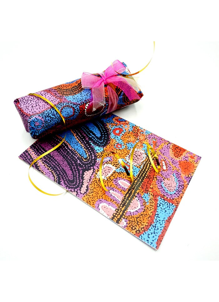 A roll of vibrant, multicolored patterned wrapping paper tied with a pink ribbon and yellow string, displayed next to a flat sheet of the same patterned paper on a white background.