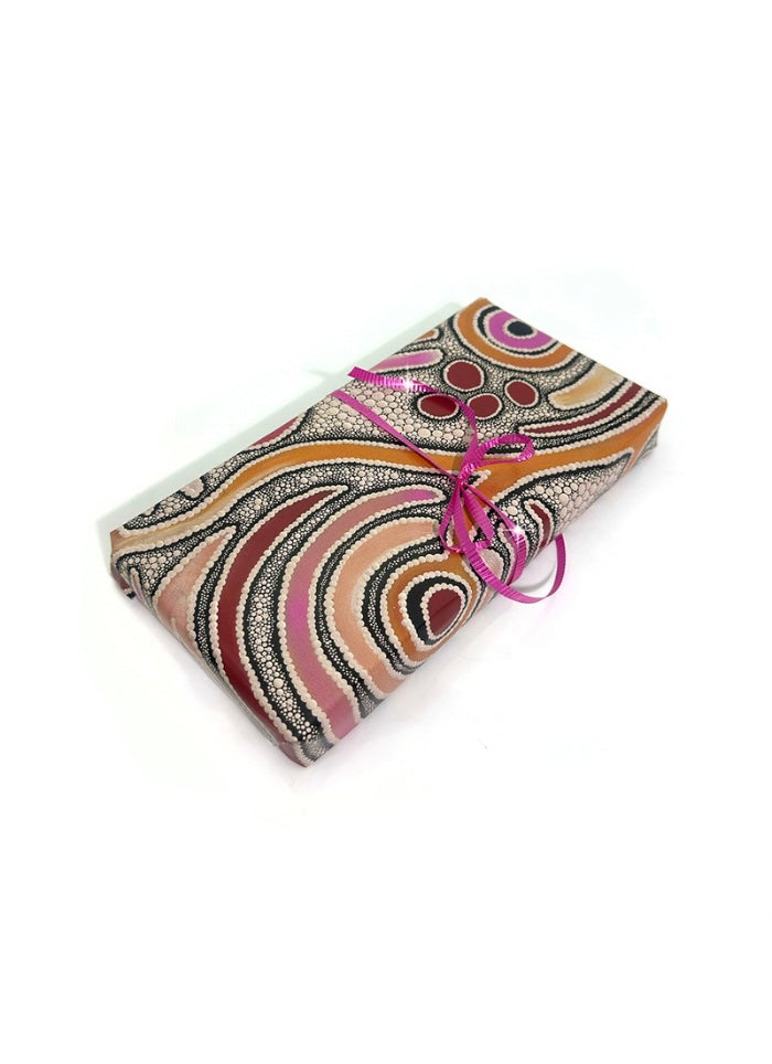 A folded sheet of wrap, featuring a vibrant Aboriginal-inspired dot art pattern in orange, pink, red, black, and white, tied with a bright pink ribbon on a white background.