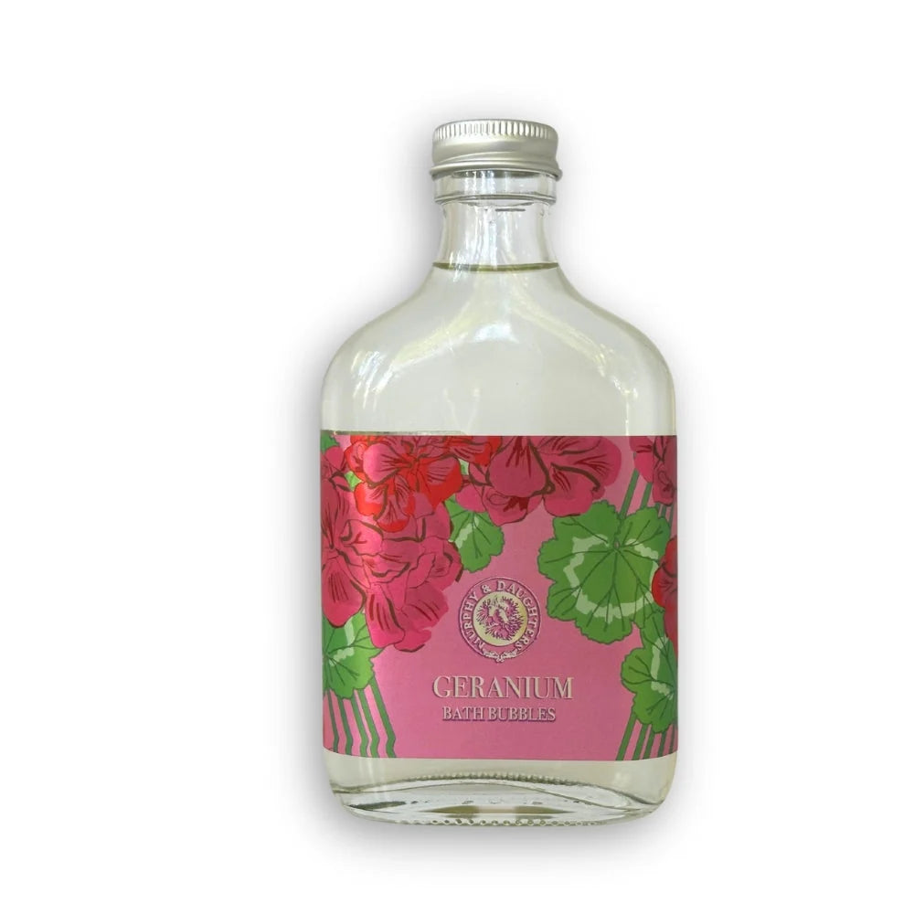 A small clear glass bottle of Bath & Body Works Geranium Bath Bubbles with a pink floral label and silver cap, isolated on a white background.