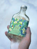 A hand holds a small clear glass bottle filled with liquid and covered in white foam, featuring a blue and green floral label that reads Green Tea and Calmsdown. The background is a soft, out-of-focus light blue and white texture.