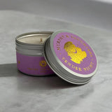 An open silver metal candle tin with a lavender purple label and gold foil lettering, next to its matching lid on a gray marble surface.