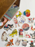 A collection of colorful, die-cut stickers featuring Japanese cultural motifs, animals, and faces, arranged on a white surface next to a wooden box.