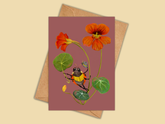 A botanical art print card featuring orange nasturtiums and a yellow-and-black ladybug on a dusty rose background, shown with a kraft paper envelope on a warm beige backdrop.