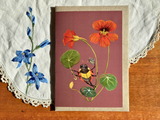 A rectangular art print or card featuring a botanical illustration of orange flowers, green leaves, and a yellow-and-black bee on a dusty mauve background, placed on a wooden surface with a white embroidered cloth underneath.