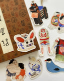 A set of colorful Japanese-style die-cut stickers arranged on a white surface next to a brown patterned box. The stickers depict traditional motifs including a rabbit mask, a human figure in red and white, a black cat, a blue bird, and a sumo wrestler.