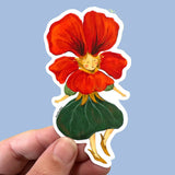 A die-cut sticker featuring a whimsical fairy-like figure with a large red flower head, gold hair, and a green leaf skirt, held by a hand against a light blue background.