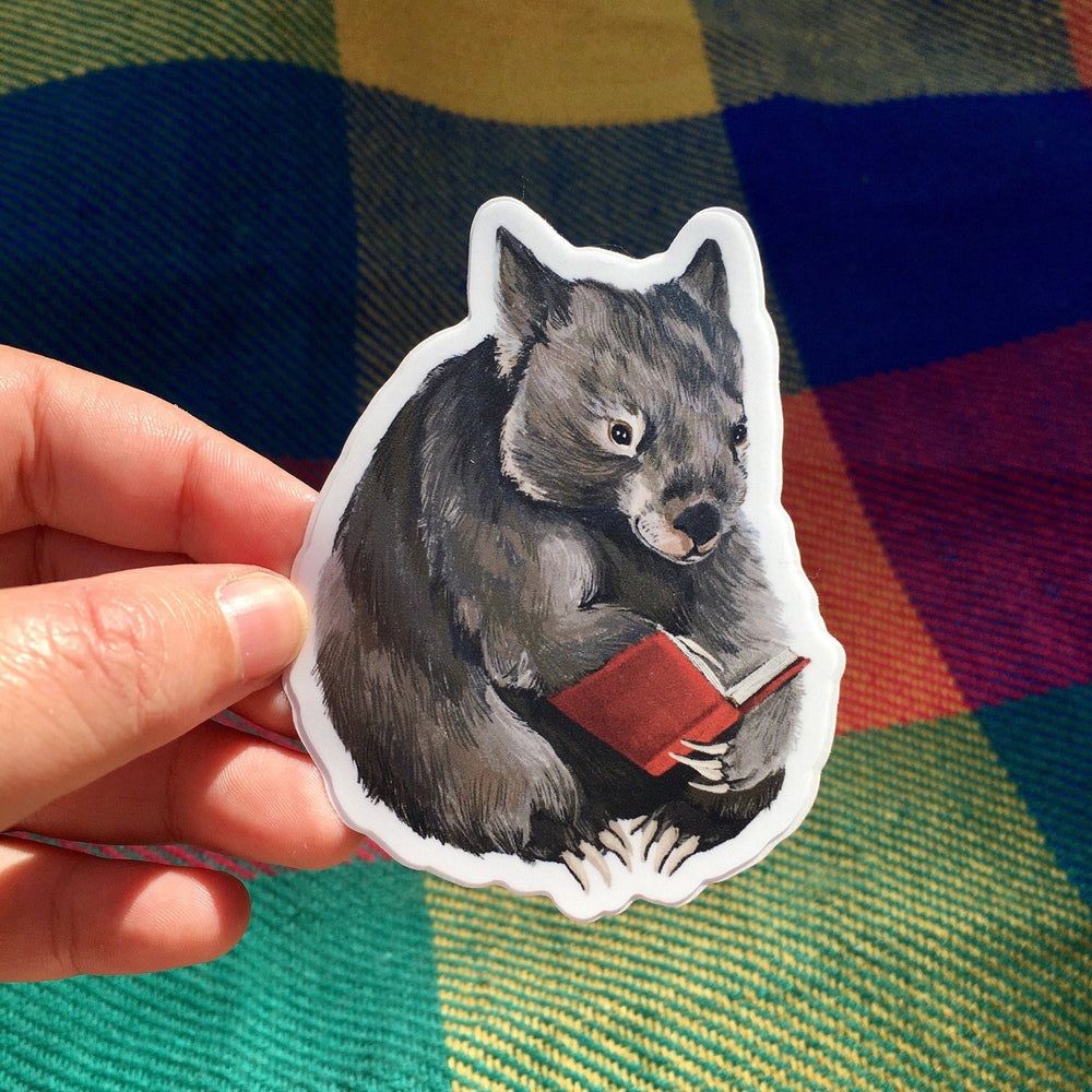 Sticker of a wombat reading a book held by a hand against a colourful fabric background