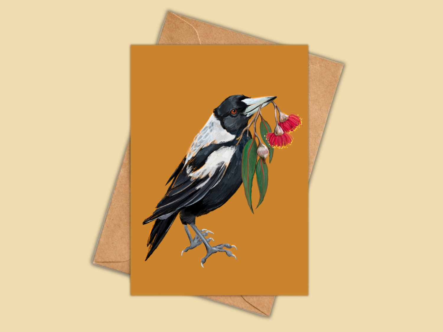 A greeting card with a warm mustard-orange background, featuring an illustration of a black-and-white bird holding red flowers, shown with a kraft envelope behind it on a light beige background.
