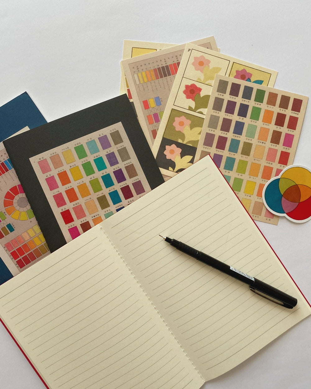 An open lined notebook with a black pen on top, surrounded by several color swatch cards and a small multicolor sticker on a white background.