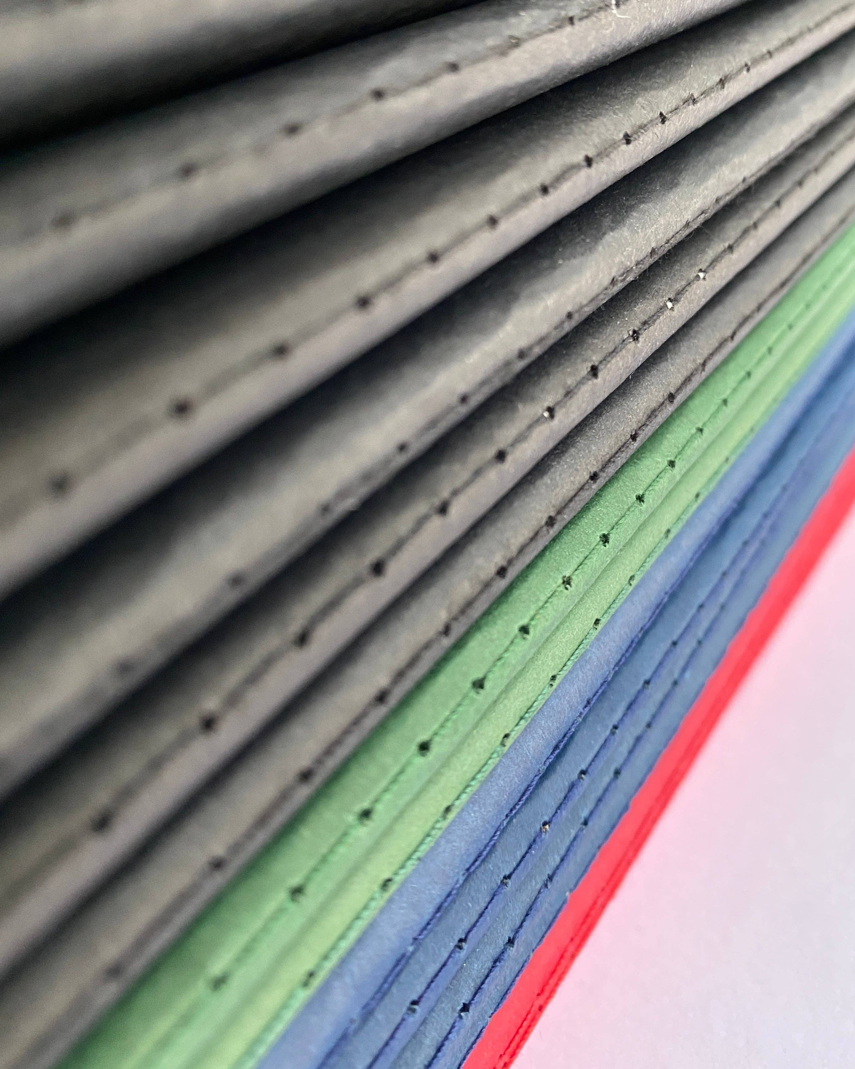 Close-up of layered fabric or textile strips in gray, green, blue, and red, stacked diagonally with visible stitched edges and small perforations.