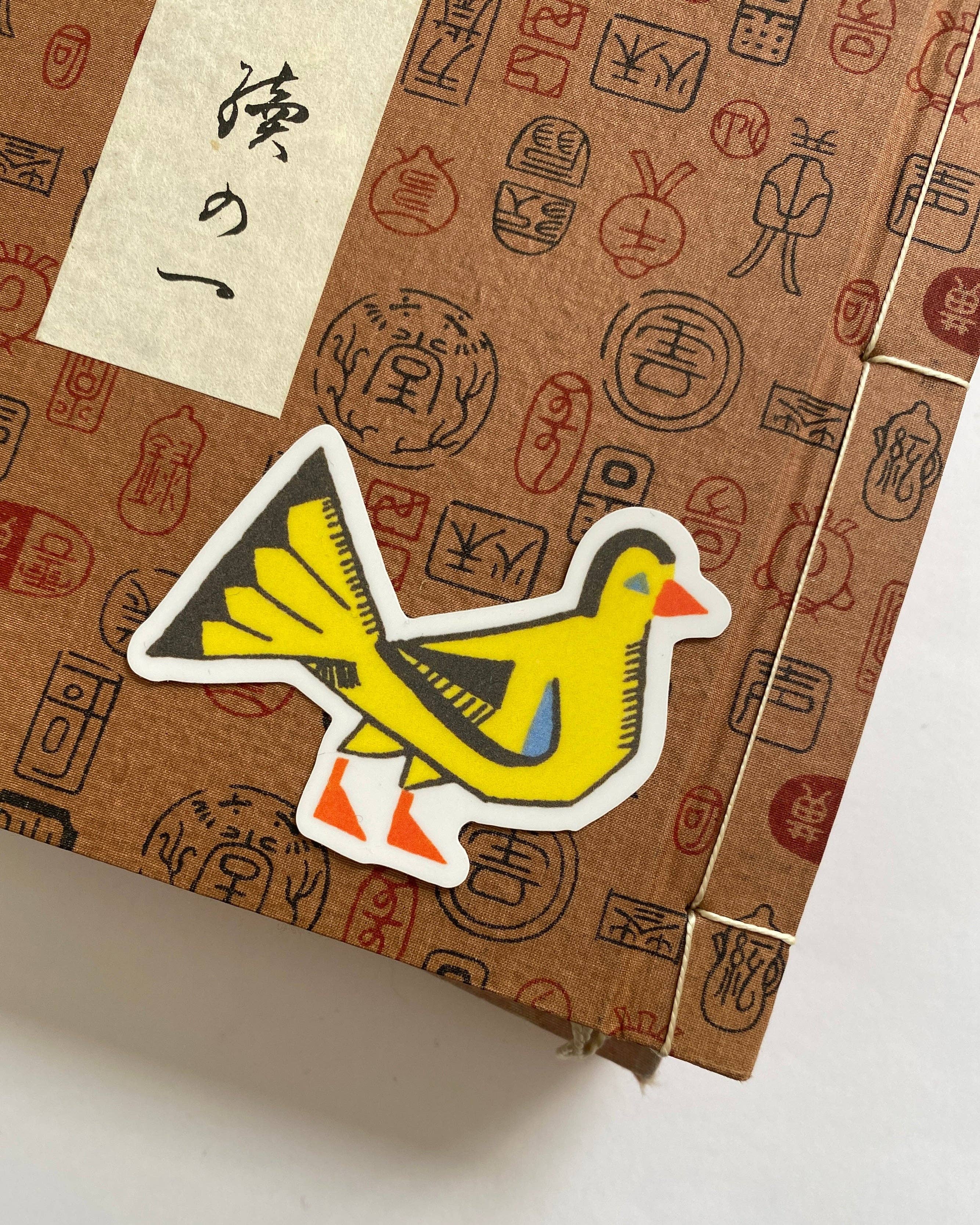 A close-up of a brown kraft-paper style box with printed red and black symbols and a cream paper label with black Japanese calligraphy. A bright yellow bird-shaped sticker with an orange beak and feet is placed on top of the box.