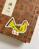 A close-up of a brown kraft-paper style box with printed red and black symbols and a cream paper label with black Japanese calligraphy. A bright yellow bird-shaped sticker with an orange beak and feet is placed on top of the box.