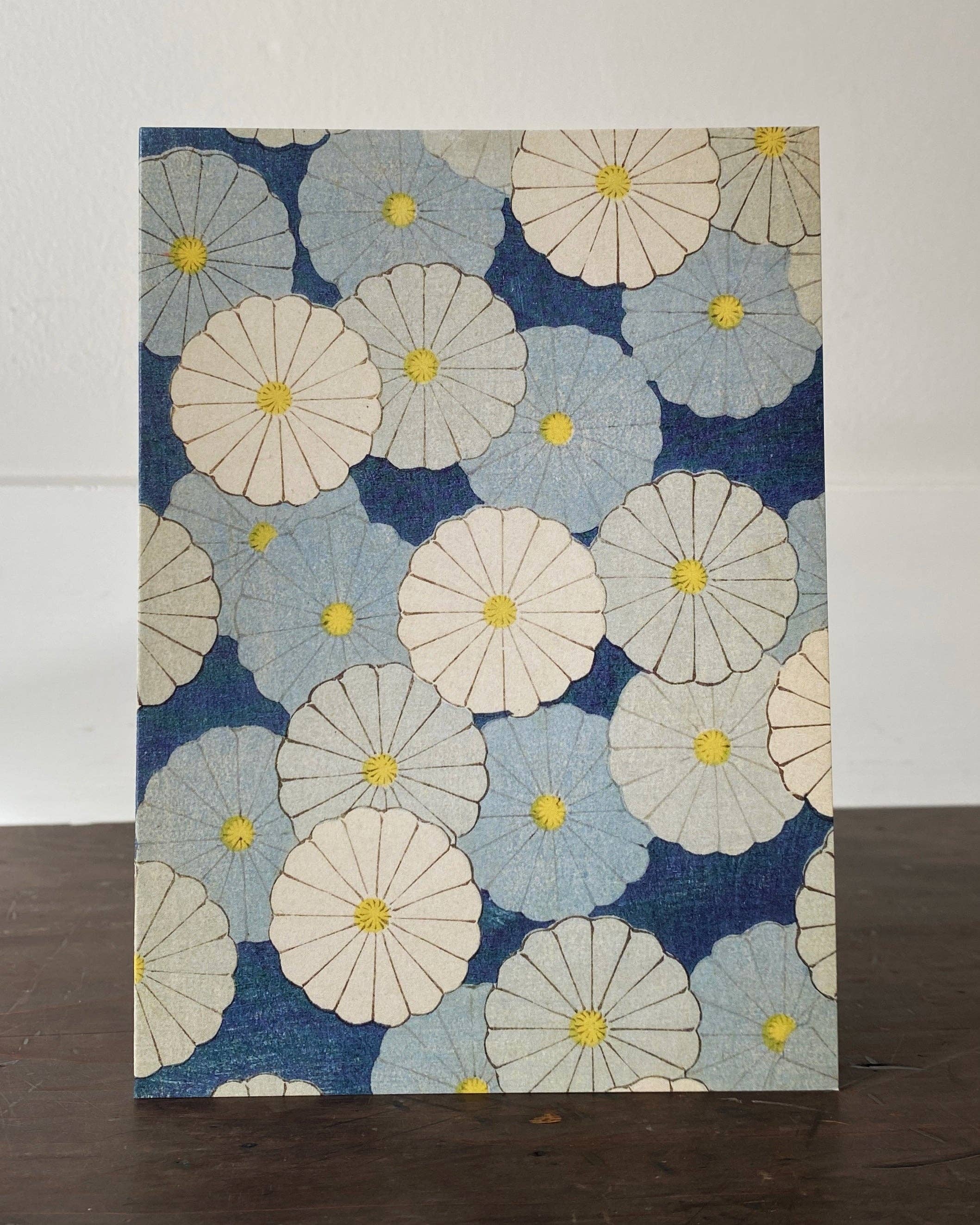 A square fabric or paper art print featuring a repeating pattern of large daisy-like flowers in cream, light blue, and navy blue with yellow centers, standing on a dark wooden surface against a white wall.