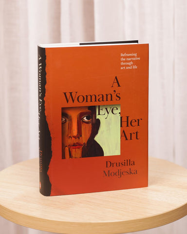Book titled 'A Woman's Eye, Her Art' by Drusilla Modjeska on a wooden surface with a white curtain background.
