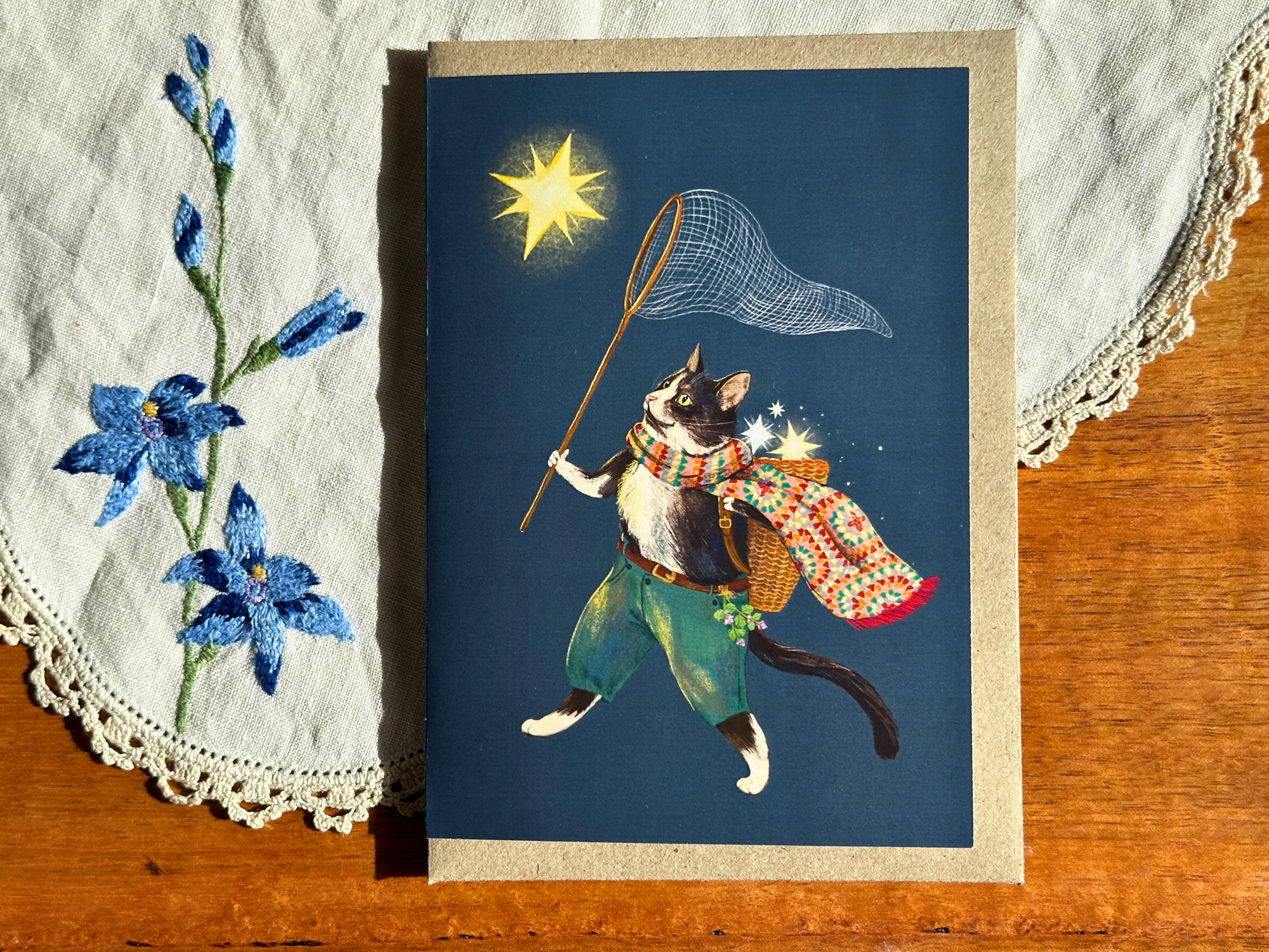 Greeting card with a cat character holding a star, surrounded by blue flowers on a textured surface.