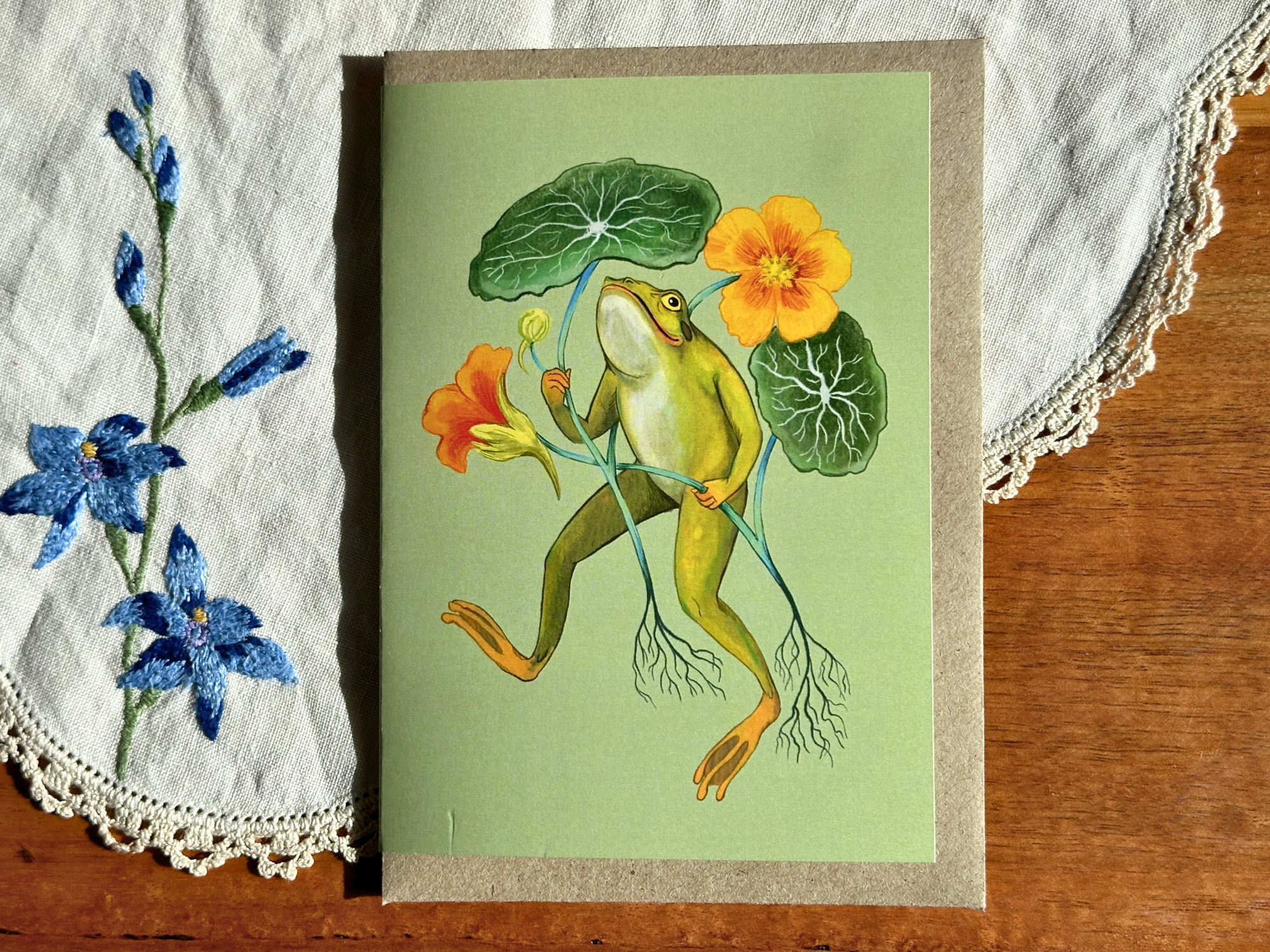 Framed artwork of a frog among flowers on a wooden surface with lace fabric.