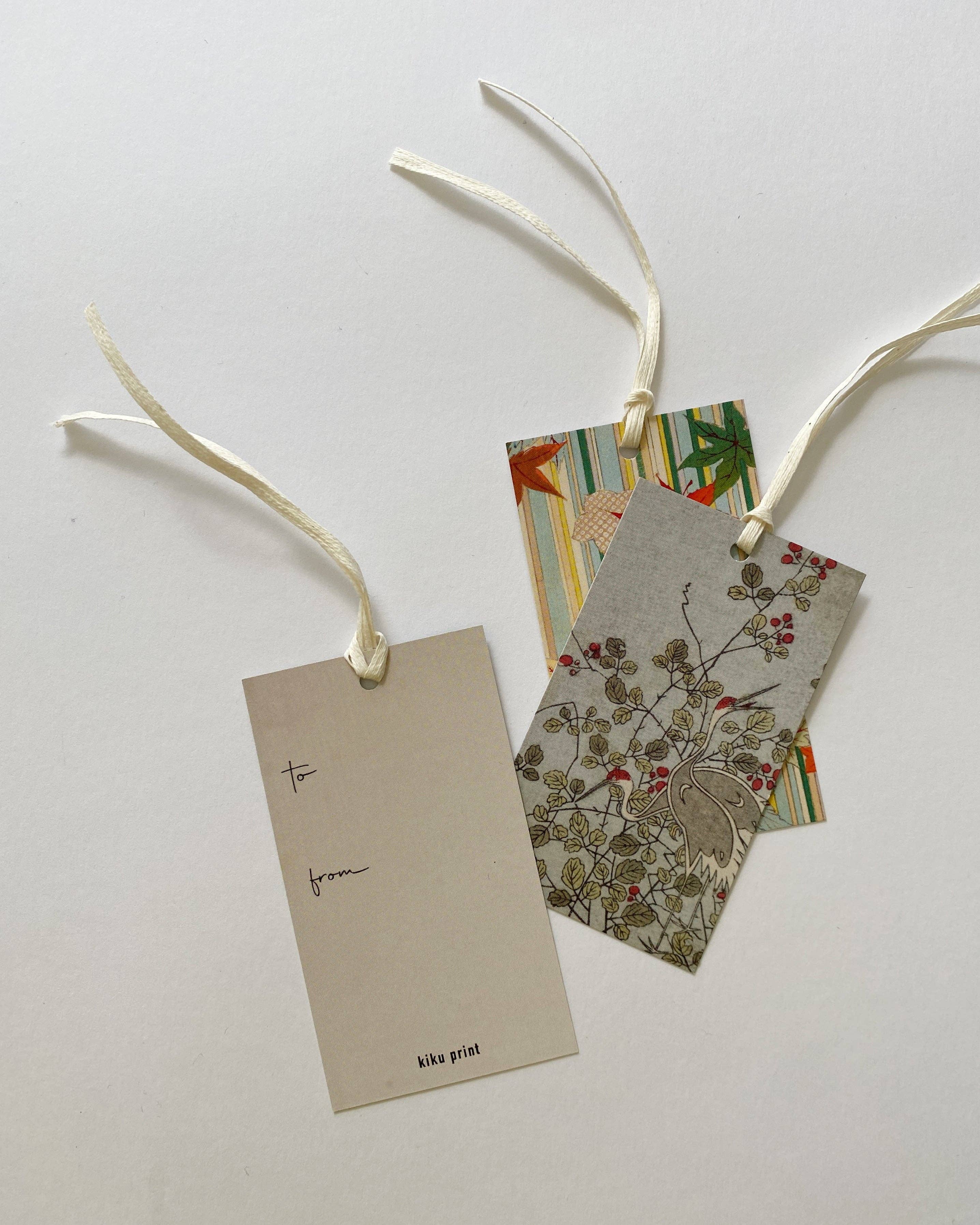 Three decorative gift tags with cream ribbons on a light gray background. One blank tag is labeled 'to from' and 'kika print', while two patterned tags behind them show floral and striped designs.