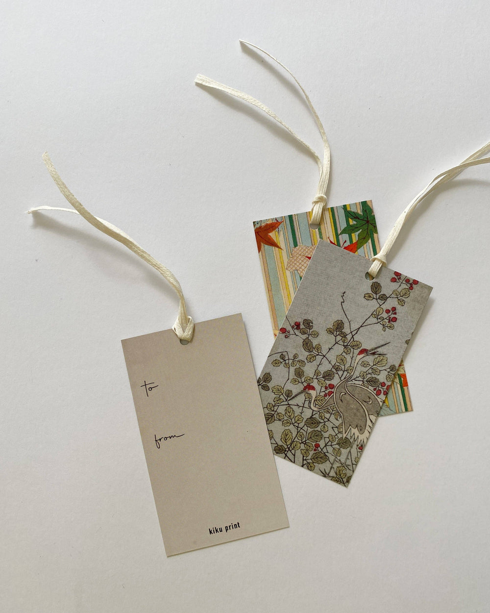 Three decorative gift tags with cream ribbons on a light gray background. One blank tag is labeled 'to from' and 'kika print', while two patterned tags behind them show floral and striped designs.