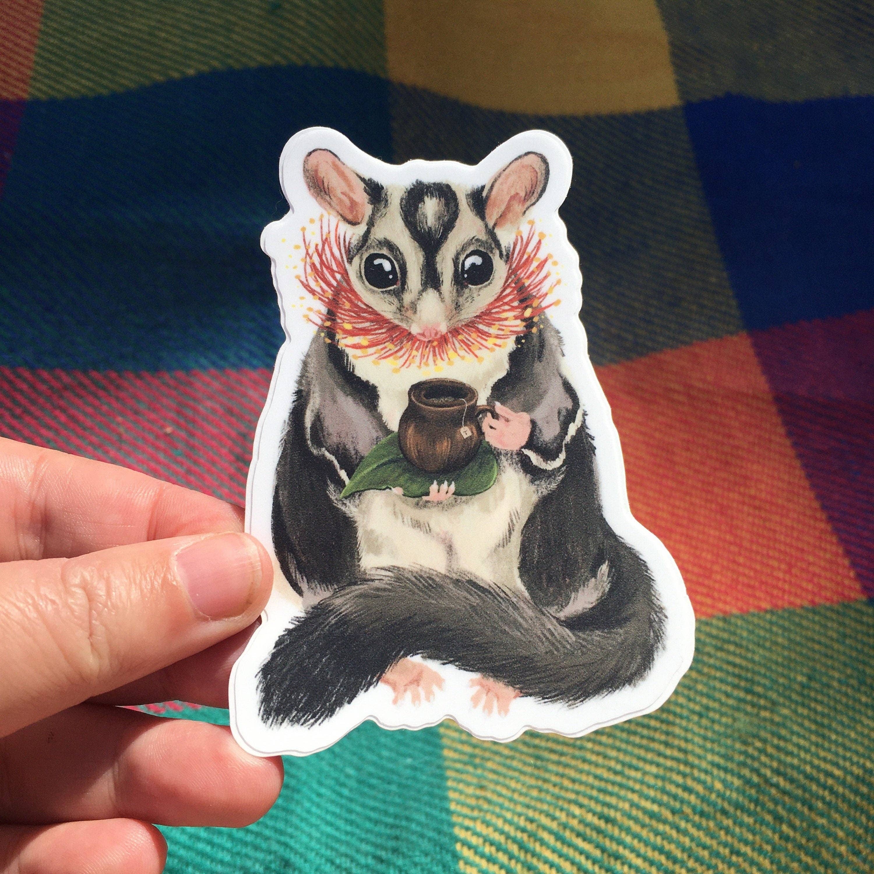 Sticker of a sugar glider holding a teacup on a checkered fabric background