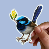 A hand holding a die-cut sticker of a blue and black fairy wren holding yellow flowers, set against a solid light blue background.