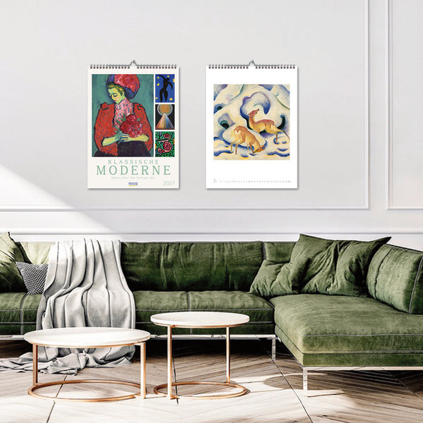 Two wall calendars featuring art on a white wall above a green sofa.