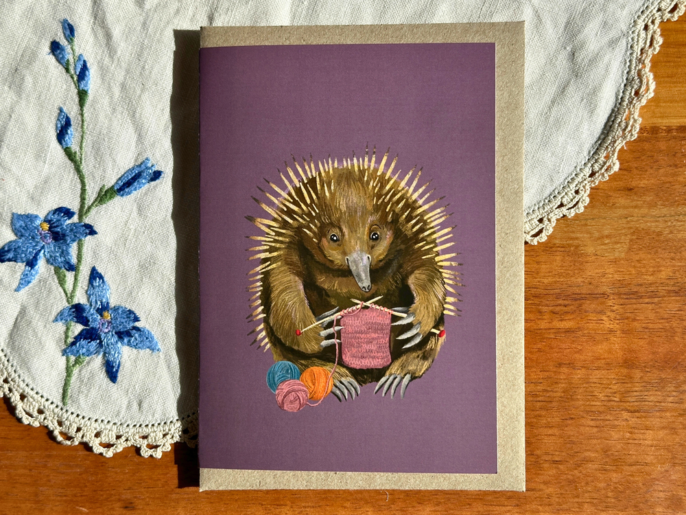 A square greeting card featuring an illustration of a brown hedgehog knitting a pink sweater, placed on a wooden table with a white embroidered cloth.