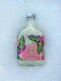 A small clear glass bottle with a silver cap and a pink decorative label reading ROSIE, placed on a bed of white foam or ice. The label features green, purple, and pink abstract floral artwork.