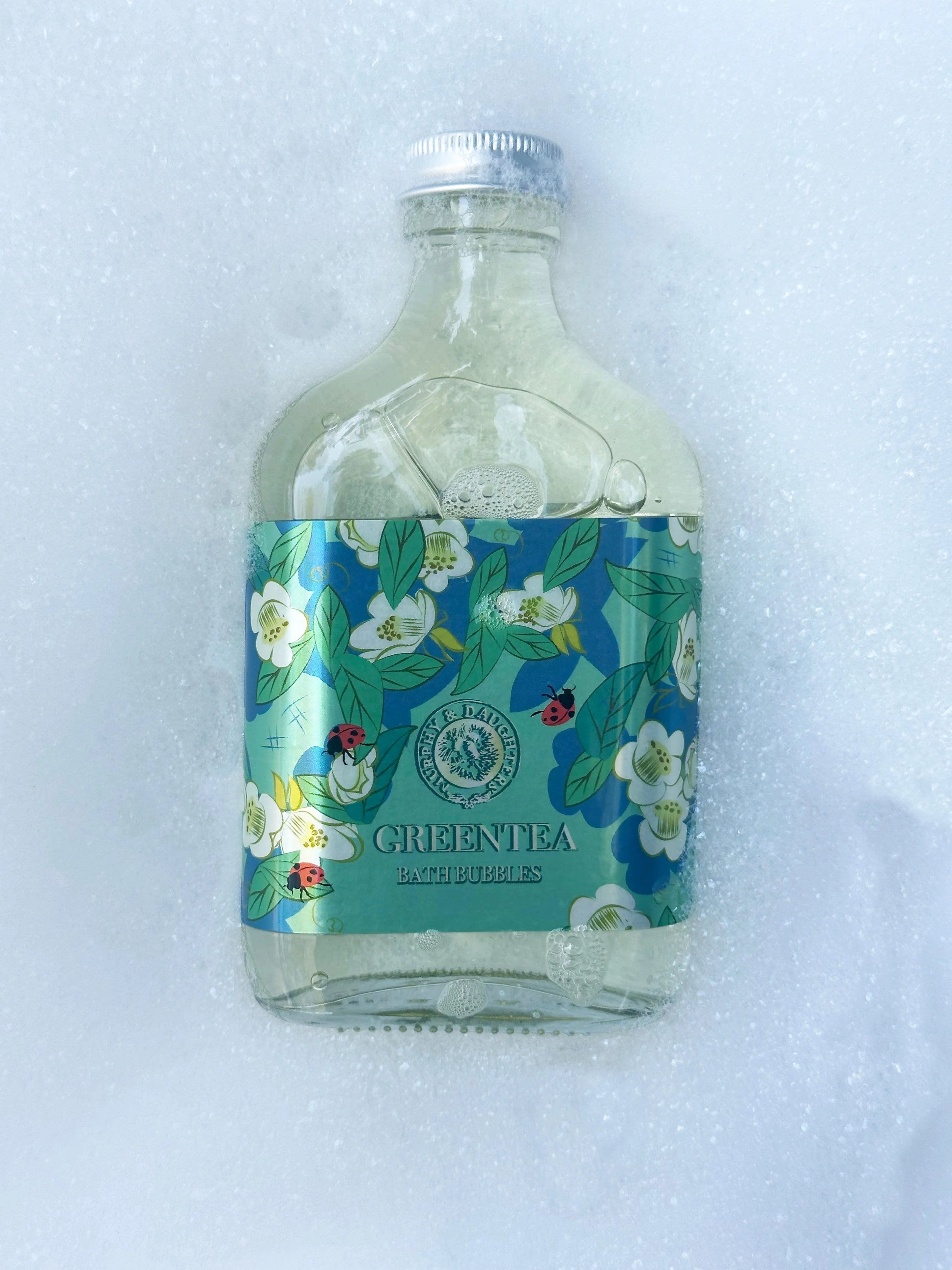 A clear glass bottle of Green Tea Cold Bubbles drink with a colorful floral label, resting on a bed of white foam.