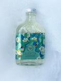 A clear glass bottle of Green Tea Cold Bubbles drink with a colorful floral label, resting on a bed of white foam.