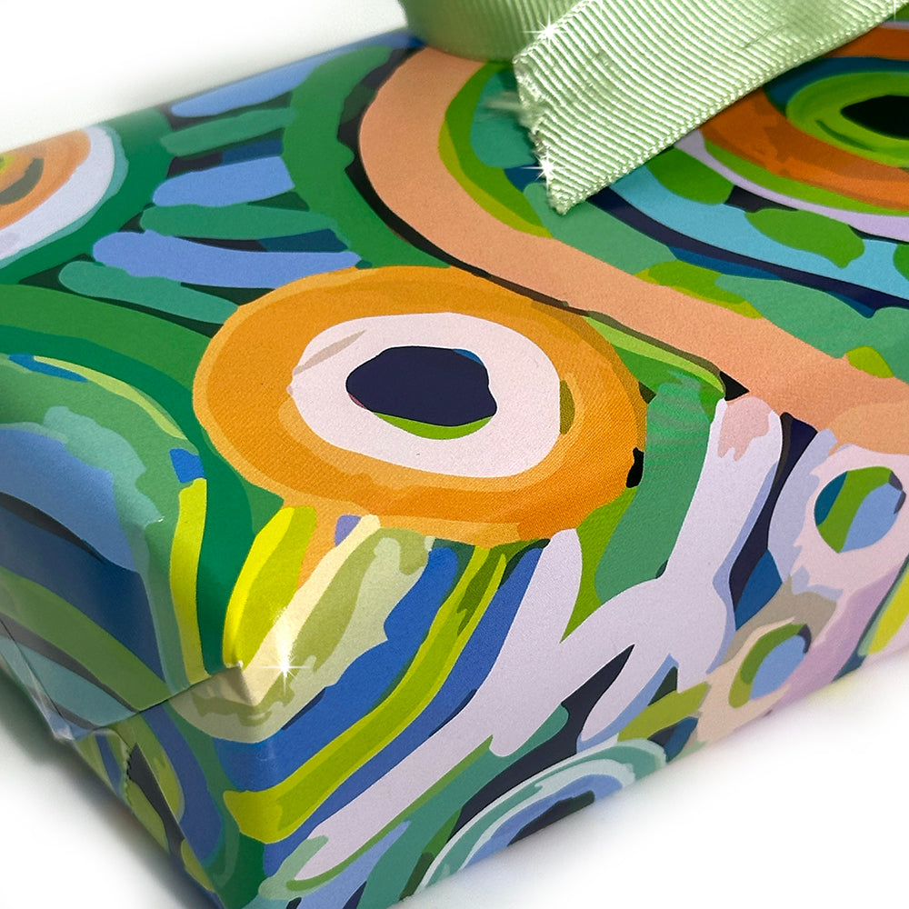 A close-up of a gift box wrapped in colorful abstract patterned paper with a light green ribbon bow, photographed on a white background.