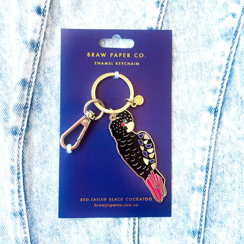Enamel keychain of a red-tailed black cockatoo on a Braw Paper Co. card against a denim background