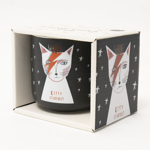 A black mug with white stars and a cat with  a lightning mark on its face says Kitty Stardust - art by Niaski.