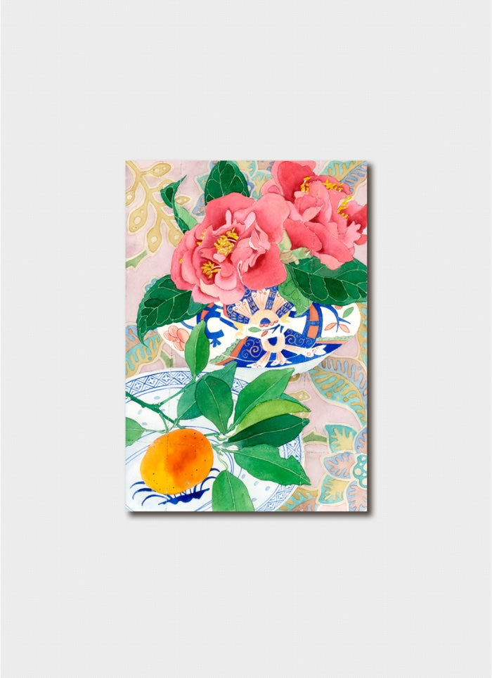 Pink camelias in a patterned bowl next to patterned plate with an orange.