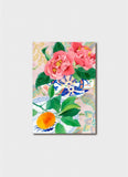 Pink camelias in a patterned bowl next to patterned plate with an orange.