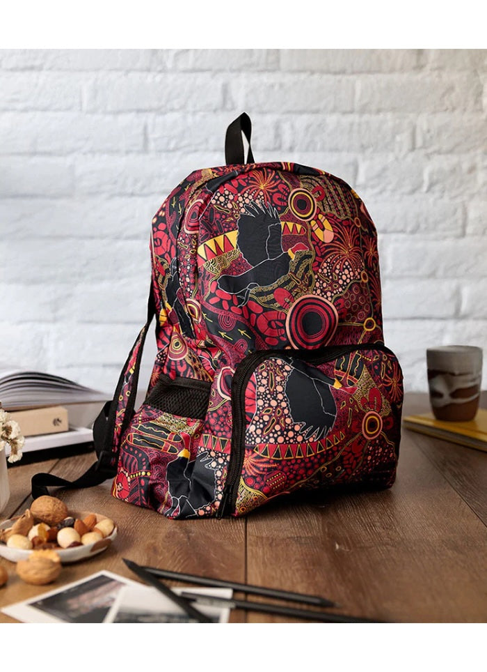 A backpack with a vibrant, dense Aboriginal-style dot art pattern in red, black, yellow, and white, displayed on a wooden table with lifestyle props against a white brick wall background.
