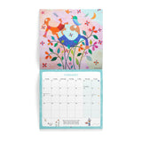 A hanging wall calendar shown open to the February page, featuring a colorful whimsical illustration on the cover with two birds, flowers, and a crescent moon above a monthly calendar grid.
