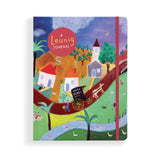 A hardcover Leunig Journal featuring a colorful, whimsical folk-art style illustration of a village scene with houses, trees, a river, and a red elastic closure band.