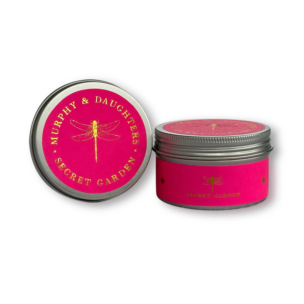 Two round metal tins for candles on a white background. One tin is open, showing a bright pink lid with gold lettering that reads 'MURPHY & DAUGHTERS SECRET GARDEN' and a gold dragonfly.
