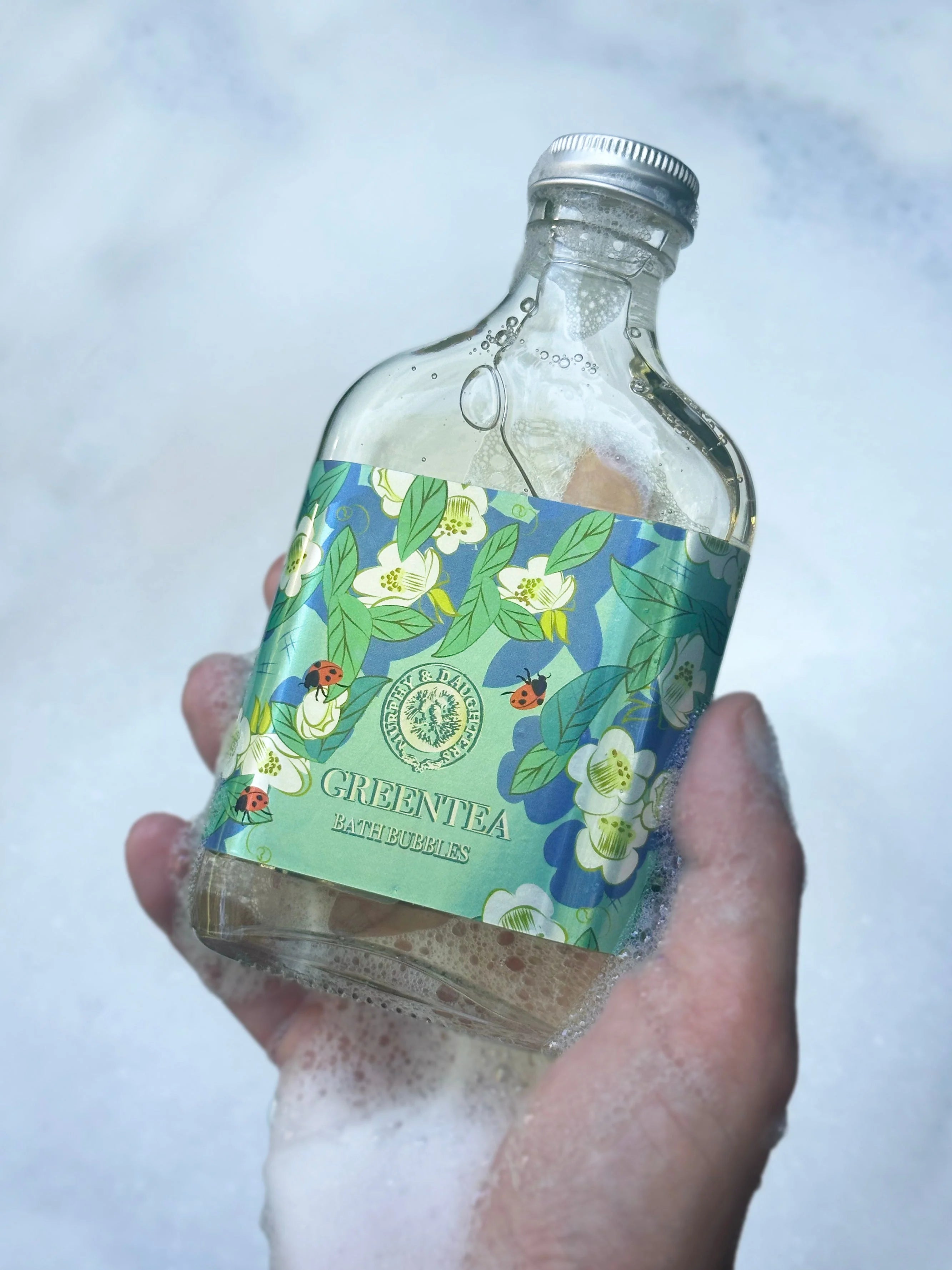 A hand holds a small clear glass bottle filled with liquid and covered in white foam, featuring a blue and green floral label that reads Green Tea and Calmsdown. The background is a soft, out-of-focus light blue and white texture.