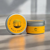 Two silver metal tins for Murely & Daughters Wild Things, one standing upright with a yellow label and one lying on its side showing the lid with a yellow palm tree design, set on a wooden surface with soft window shadows.