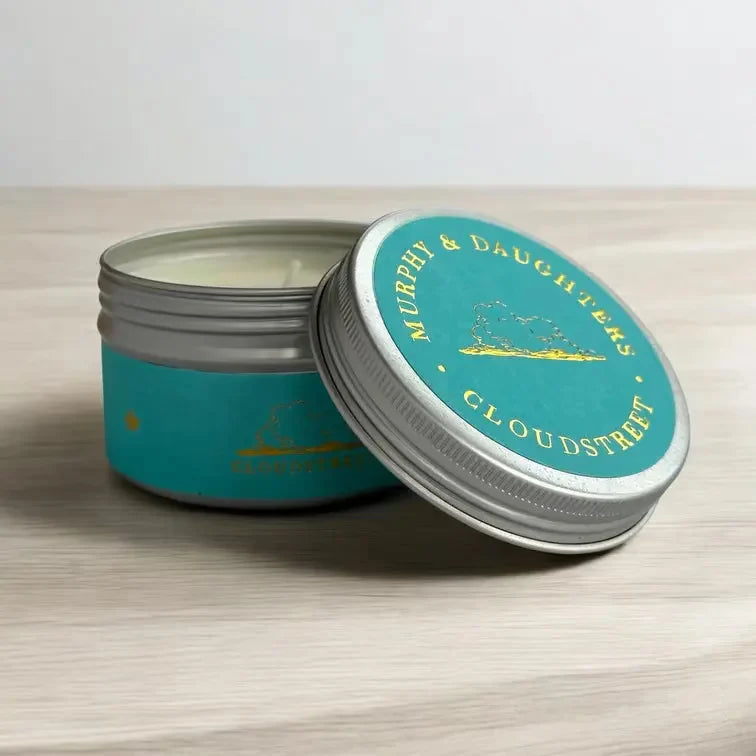 An open silver metal tin of candle with a turquoise label and matching lid leaning against it on a light wood surface. The lid reads 'MURPHY & DAUGHTERS CLOUDSTREET' in gold lettering.