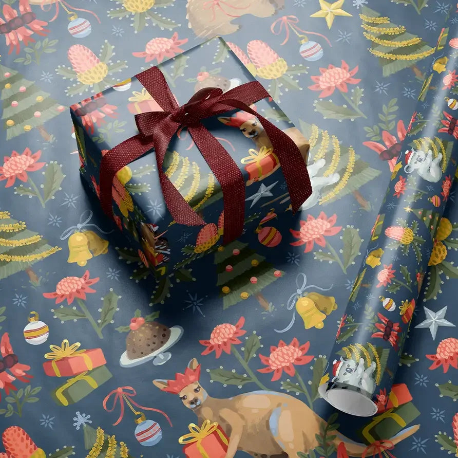 Holiday-themed wrapping paper with a festive print, shown wrapped around a gift box tied with a dark red ribbon and a matching rolled sheet on a blue background. The pattern features Christmas trees, ornaments, bells, stars, and a dog wearing a red Santa hat.