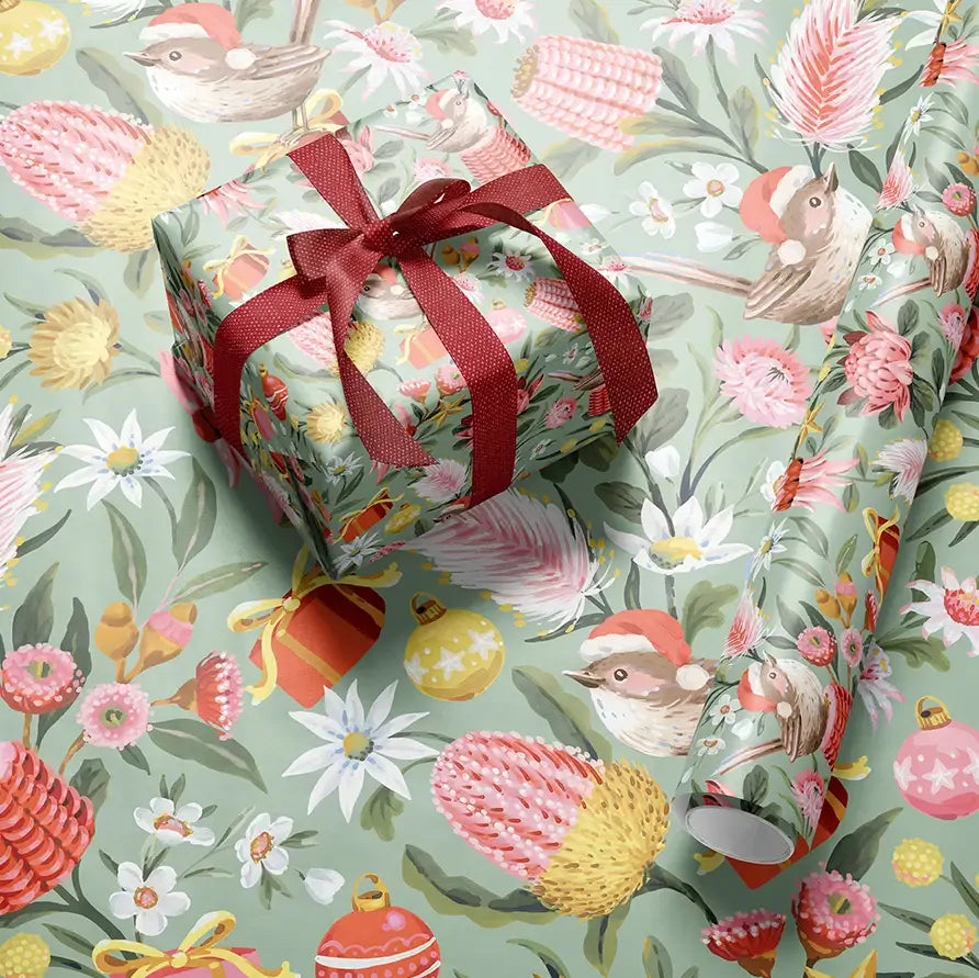 A wrapped gift box tied with a dark red ribbon sits on top of matching wrapping paper printed with a whimsical botanical and bird pattern. The design features small birds, pink flowers, yellow and red botanical elements, and ornaments on a pale sage green background.